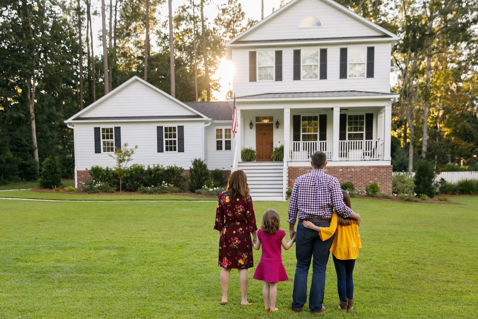 Family from behind standing outside front of new construction white siding farmhouse in the suburbs